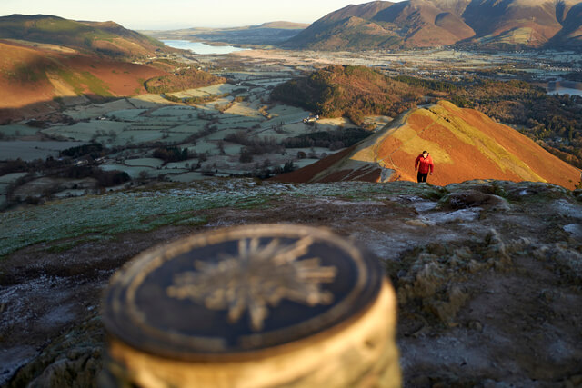 A hiker approaching the mountain summit marker at Catbells
