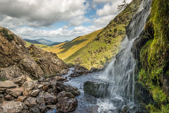 A waterfall cascading down a mossy rockface with countryside in the background