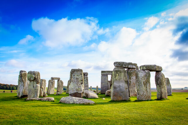 A view of stone henge sitting on the grass with the clouds in the sky above
